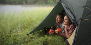 Een foto van een jong stel dat samen uit een tent kijkt op de groene oever van een meer. Ze hebben oranje kampeerbekers vast. Staatsbosbeheer sluit alle wildkampeerterreinen.