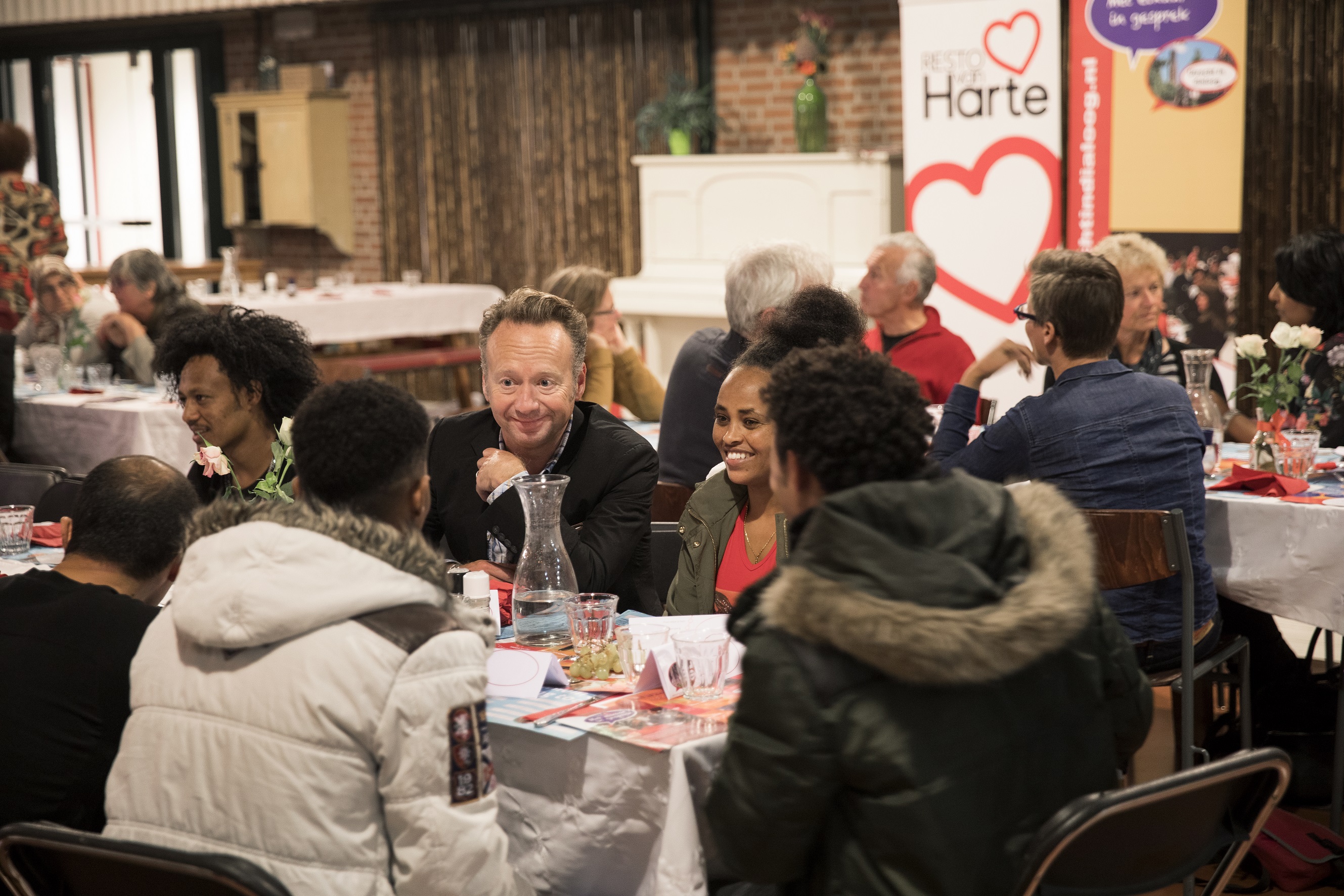 Op deze foto is Joris Linssen te zien, hij zit met een groepje mensen aan tafel in een restaurant van Resto VanHarte